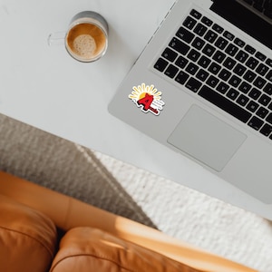 May include: A sticker featuring a large red letter A with a small red bow on top, positioned in front of a yellow sunburst graphic. The text reads "Annie" in black letters below the A, with smaller text reading "The Sun Will Come Out Tomorrow".