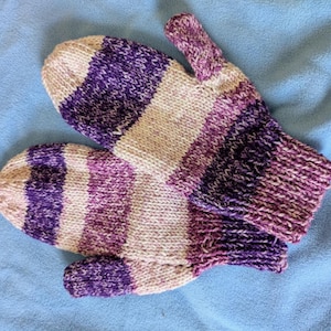 Pode incluir: Um par de luvas de malha listradas em roxo, branco e bege. As luvas estão sobre uma superfície azul. As luvas são projetadas para manter as mãos aquecidas. Medem cerca de 20 cm de comprimento.