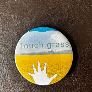 May include: A round button with a blue sky and white clouds, a field of yellow flowers, and a white hand reaching up. The text "Touch grass" is printed in green letters.