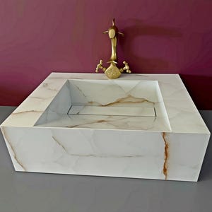 May include: A square white sink with brown veining patterns throughout the stone surface. The basin features a flat rectangular drain cover in the center. A gold-toned metal faucet with two handles is positioned directly behind the sink basin.