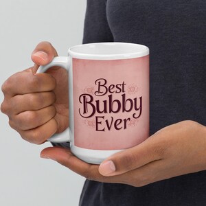 May include: White ceramic mug with the text "Best Bubby Ever" in a decorative script. The mug has a pink background with a white rim and handle. The mug is held by a person.