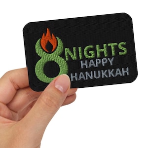 Könnte beinhalten: Schwarzer rechteckiger Aufnäher mit den Worten "8 NIGHTS HAPPY HANUKKAH" in Grün und Grau. Die Zahl 8 ist grün mit einer orangefarbenen Flamme darüber. Der Aufnäher wird von einer Hand gehalten.