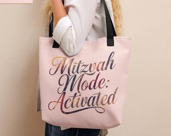 Könnte beinhalten: Hellrosa Tragetasche mit schwarzen Henkeln. Auf der Tasche steht in dekorativer Schrift der Text "Mitzvah Mode: Activated". Der Text hat ein farbenfrohes, galaxieähnliches Design.