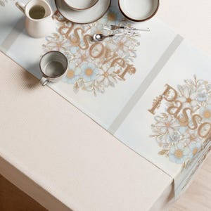 May include: A light blue and white table runner with floral designs and the word "Passover" in gold lettering. The runner features a repeating pattern of flowers and is set with tableware.