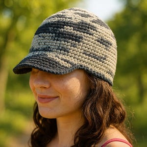May include: A crocheted hat with a brim, featuring a camouflage pattern in shades of gray, black, and beige. The hat is worn by a person with brown hair and a pink tank top, set against a blurred green background.