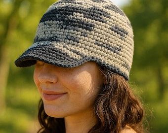 May include: A crocheted hat with a brim, featuring a camouflage pattern in shades of gray, black, and beige. The hat is worn by a person with brown hair and a pink tank top, set against a blurred green background.