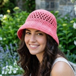 Può includere: Un cappello a secchiello rosa all'uncinetto con una tesa larga. Il cappello ha un design a trama aperta ed è indossato da una persona con lunghi capelli scuri. Lo sfondo presenta una vegetazione lussureggiante e fiori, creando un ambiente naturale.