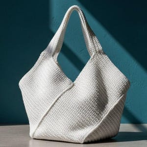 May include: A white, textured tote bag with a unique geometric design. The bag has a single handle and is made of a woven material. The bag is set against a teal blue wall with a shadow.