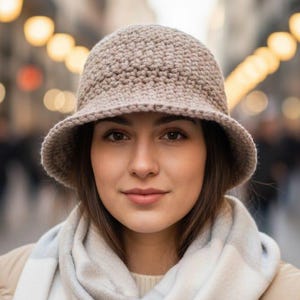 Può includere: Un cappello a secchiello all'uncinetto beige con una tesa larga. Il cappello è indossato con una sciarpa e una giacca di colore chiaro. L'immagine è scattata all'aperto, con uno sfondo sfocato di una via cittadina.