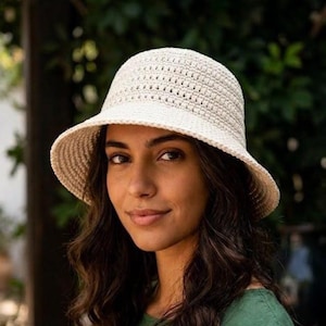 Può includere: Un cappello da sole all'uncinetto color crema con tesa larga. Il cappello è indossato da una persona con capelli scuri e un top verde. Lo sfondo presenta una vegetazione lussureggiante e vasi di ceramica.
