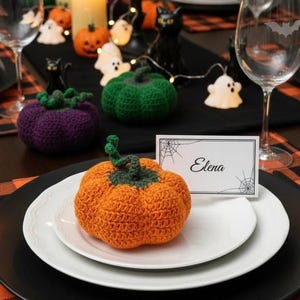 May include: A crocheted orange pumpkin with a green stem sits on a white plate atop a black charger. A white place card with the name Elena and spiderweb graphics is positioned next to the pumpkin. The table features orange and black plaid mats.