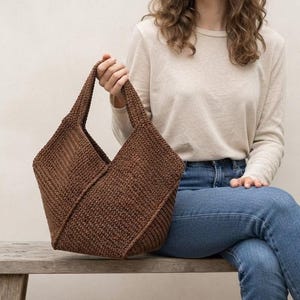 May include: A dark brown, hand-crocheted tote bag featuring a structured, angular silhouette with a wide opening and two integrated handles. The bag is crafted from a textured, woven yarn material with visible stitch patterns throughout the exterior.
