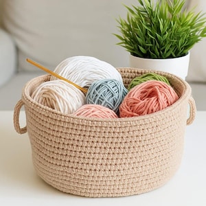 以下が含まれることがあります： 白、青、ピンク、緑色の毛糸玉が入ったベージュ色の編みかご。金色の編み棒がバスケットの中にあります。右側には小さな鉢植えの植物があります。