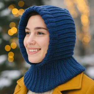 Puede incluir: Primer plano de un pasamontañas de punto azul. El pasamontañas está hecho de una tela de punto grueso y acanalado. La persona lleva un abrigo amarillo y sonríe. El fondo está borroso con luces bokeh.