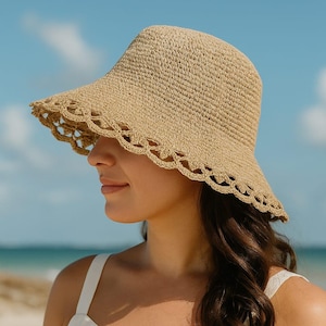 Può includere: Un cappello da sole beige intrecciato con un bordo smerlato. Il cappello è indossato da una persona su una spiaggia, con l'oceano e il cielo sullo sfondo. La tesa del cappello è larga circa 10 cm.