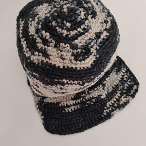 Un cappellino da baseball all'uncinetto fatto a mano nei toni del blu scuro, grigio e crema. Il cappellino è poggiato su un tavolo bianco ed è visto dall'alto. Un accessorio sportivo e unisex.