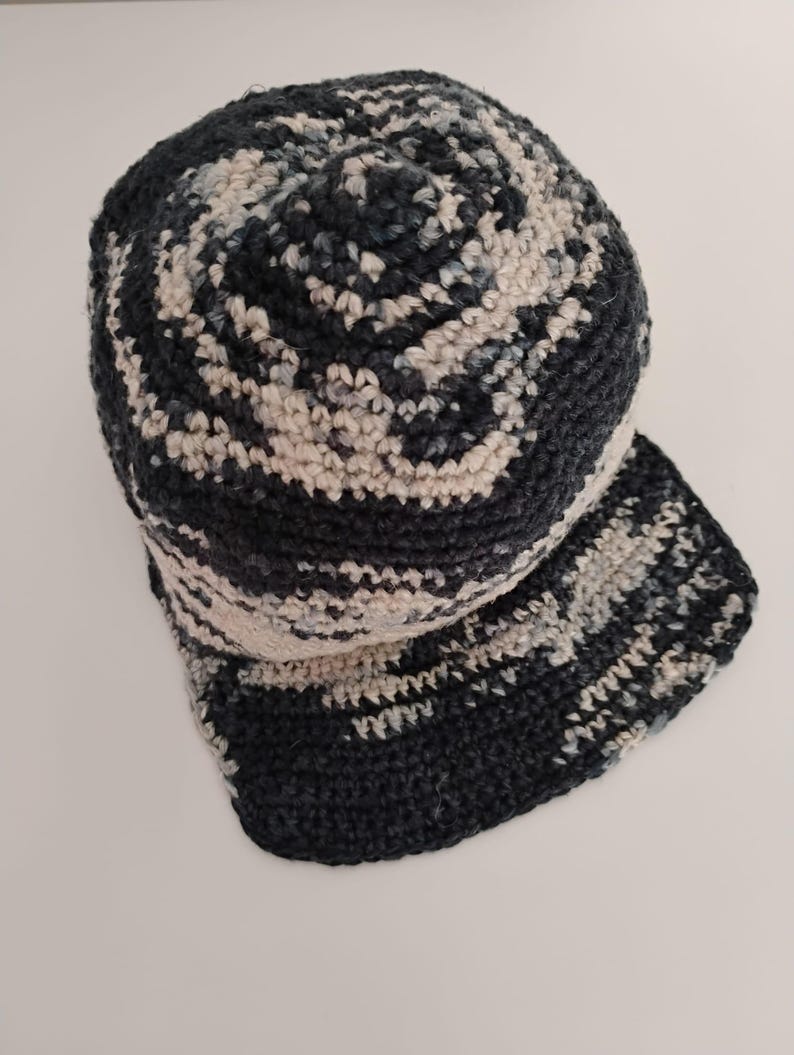 Un cappellino da baseball all'uncinetto fatto a mano nei toni del blu scuro, grigio e crema. Il cappellino è poggiato su un tavolo bianco ed è visto dall'alto. Un accessorio sportivo e unisex.