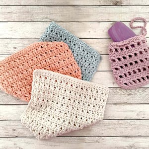 Può includere: Una collezione di articoli all'uncinetto fatti a mano in vari colori. L'immagine presenta quattro salviette in pesca, azzurro e crema, insieme a un sacchetto portasapone lavanda con una saponetta all'interno. Gli articoli sono disposti su una superficie di legno chiaro.