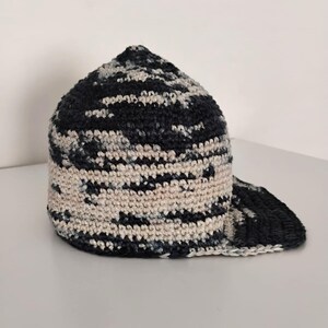 Cappello all'uncinetto con motivo mimetico blu navy e bianco sporco. Il cappello ha una corona arrotondata e una visiera curva. La trama dell'uncinetto è visibile, aggiungendo fascino artigianale.