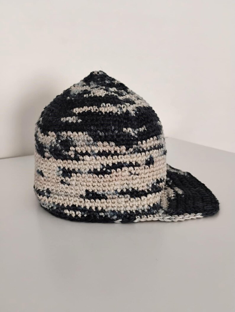 Cappello all'uncinetto con motivo mimetico blu navy e bianco sporco. Il cappello ha una corona arrotondata e una visiera curva. La trama dell'uncinetto è visibile, aggiungendo fascino artigianale.