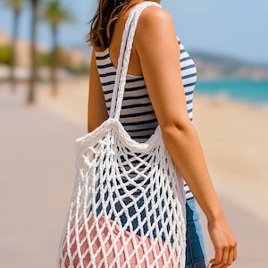 May include: A white, open-weave net bag with a pink item inside is carried over the shoulder. The bag has a braided handle. The person is wearing a striped tank top, denim shorts, and a straw hat. The background shows a beach scene.