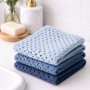 Può includere: Una pila di quattro salviette quadrate all'uncinetto in varie tonalità di blu. La salvietta superiore è azzurra, seguita da blu medi e scuri, creando un effetto sfumato. Le salviette hanno un design a trama aperta.