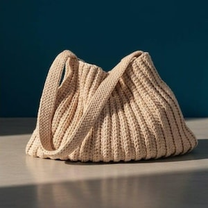 May include: A beige, hand-crocheted tote bag with a ribbed texture. The bag has a rounded bottom and two handles. The bag is sitting on a light-colored surface with a dark blue background.