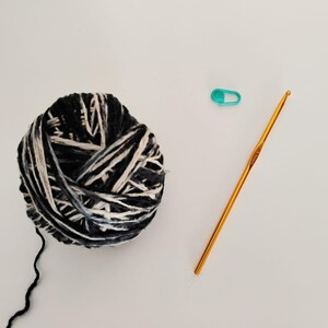 Un gomitolo di filato in lino variegato bianco e blu navy con un filo che si estende da esso. Sono presenti anche un uncinetto dorato e un segnapunti turchese, il tutto su una superficie bianca. Ideale per progetti di artigianato.