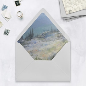 May include: A white envelope with a landscape painting on the inside. The painting depicts a mountain scene with trees, in shades of blue, green, and white. A ring and stamps are scattered around the envelope.