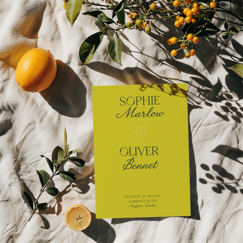 May include: A bright yellow invitation with the names Sophie Marlow and Oliver Bennet in elegant script. The invitation is surrounded by lemons, greenery, and berries on a white cloth.