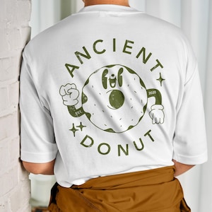 Könnte beinhalten: Weißes T-Shirt mit einer grünen Grafik eines Donuts mit Augen und Armen. Der Donut hat den Text "ANCIENT DONUT" darum herum und "EST. 2020" über dem Donut.