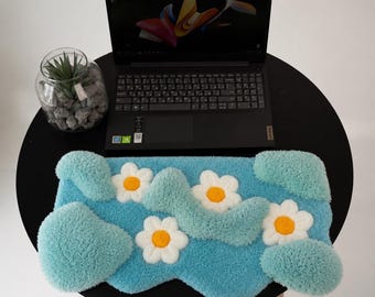 May include: A rectangular tufted rug featuring a light blue background with three white daisy flowers with yellow centers and raised, textured teal-colored cloud-like shapes. The rug is positioned in front of a black laptop on a round black table.