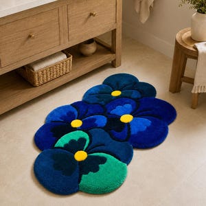 May include: A tufted floor rug shaped like a cluster of pansy flowers. The rug features overlapping petals in shades of deep navy blue, royal blue, and bright teal, with a small, circular, vibrant yellow center in each of the three flower shapes.