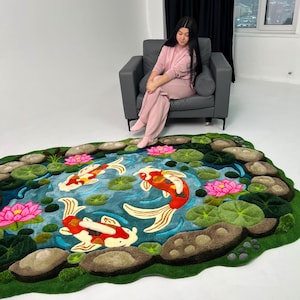 May include: A rectangular rug featuring a pond scene with three koi fish in shades of red, white, and orange. The design includes vibrant pink lotus flowers, green lily pads, and textured brown stones bordering the blue water surface.
