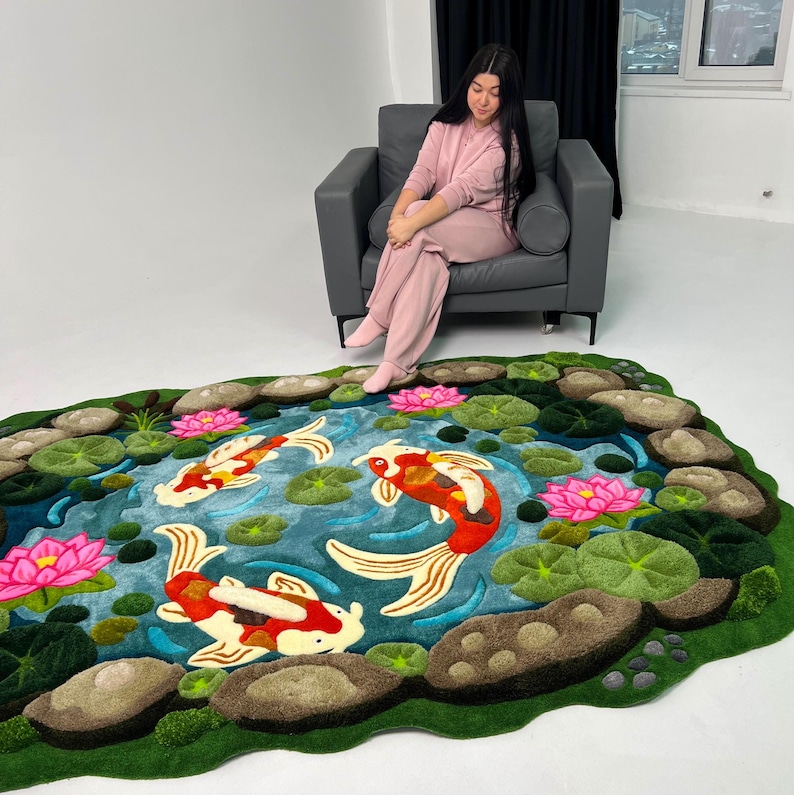 May include: A rectangular rug featuring a pond scene with three koi fish in shades of red, white, and orange. The design includes vibrant pink lotus flowers, green lily pads, and textured brown stones bordering the blue water surface.
