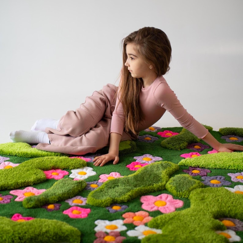 May include: A plush, tufted rug featuring a vibrant green moss-like texture interspersed with colorful, raised flower motifs in shades of pink, white, purple, and orange. The rug has an irregular, organic shape with soft, textured surfaces.