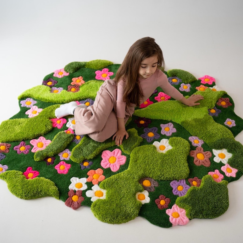 May include: A tufted area rug with an irregular, organic shape featuring a deep green background, raised light green moss-like patches, and various colorful flowers in shades of pink, orange, white, and purple with yellow centers.