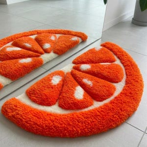 Op de afbeelding: Een halfrond getuft tapijt in de vorm van een sinaasappelschijfje. Het tapijt heeft een dikke, getextureerde oranje rand en binnensegmenten in feloranje met witte accenten, die het uiterlijk van een citrusvrucht op de vloer nabootsen.