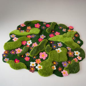 May include: A tufted rug with an irregular, organic shape featuring a deep green base. The surface is decorated with raised, textured light green moss-like patches and numerous small flowers in shades of pink, purple, white, and orange.