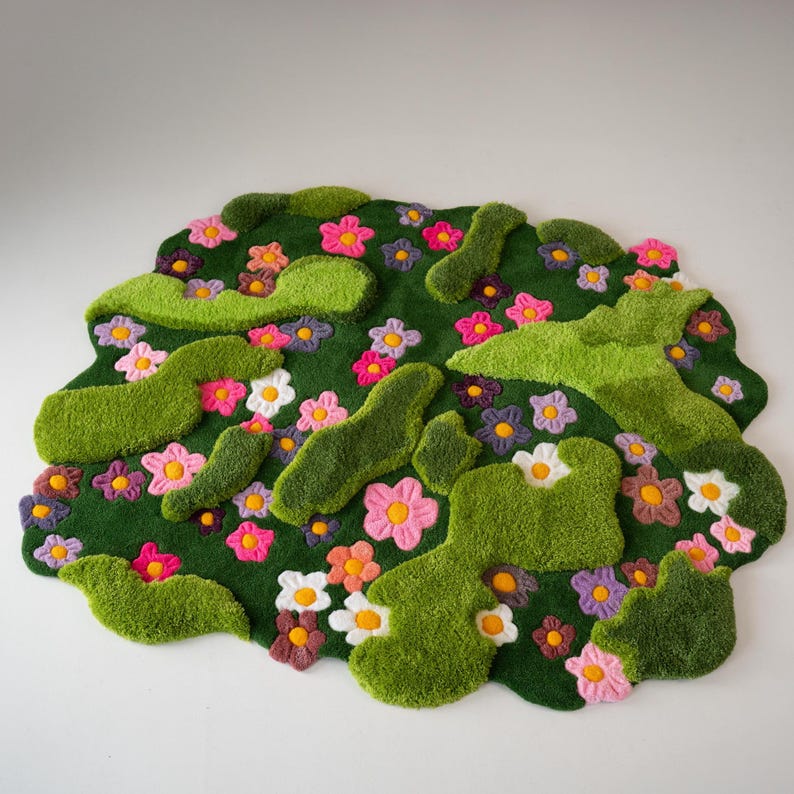 May include: A tufted rug with an irregular, organic shape featuring a deep green base. The surface is decorated with raised, textured light green moss-like patches and numerous small flowers in shades of pink, purple, white, and orange.