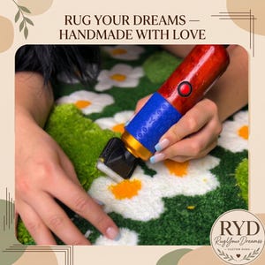 May include: A rug tufting clipper with a red handle, blue grip, and gold accent, being used to trim a green rug featuring white daisy patterns with yellow centers. The text RUG YOUR DREAMS &mdash; HANDMADE WITH LOVE is visible at the top of the frame.