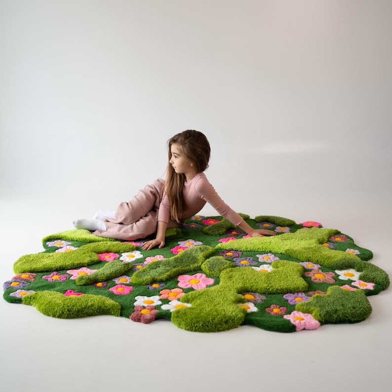 May include: A tufted rug featuring a vibrant green moss-like texture interspersed with various colorful flowers in shades of pink, white, orange, and purple. The rug has an irregular, organic shape with raised, plush sections throughout the surface.