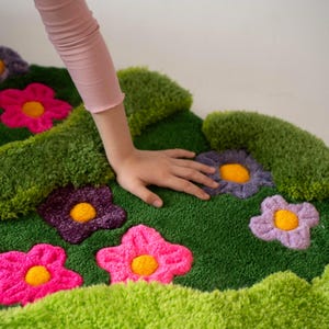 May include: A tufted rug featuring a vibrant green background with a textured, grassy appearance. The rug is decorated with several plush, multi-colored flowers in shades of bright pink, deep purple, and light lavender, each with a yellow center.