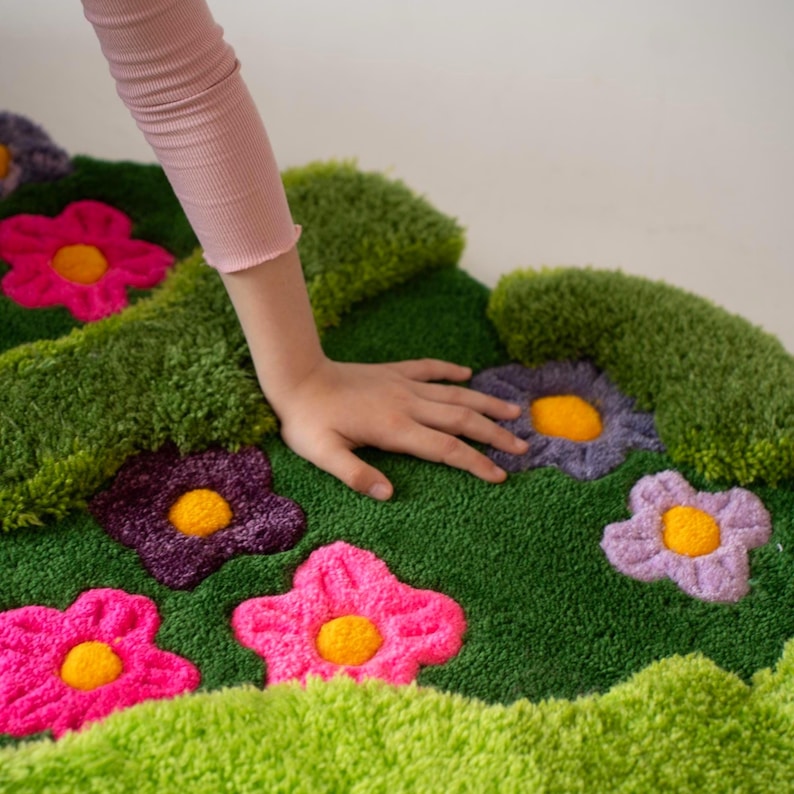 May include: A tufted rug featuring a vibrant green background with a textured, grassy appearance. The rug is decorated with several plush, multi-colored flowers in shades of bright pink, deep purple, and light lavender, each with a yellow center.