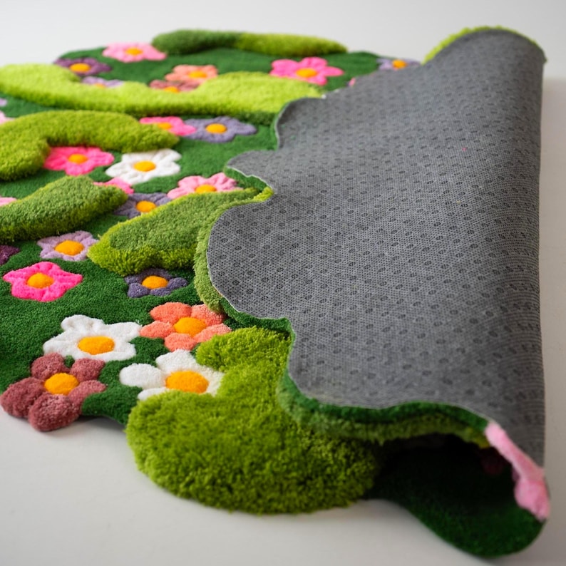 May include: A tufted rug featuring a vibrant green background with raised, textured lime green shapes and a variety of colorful flowers in pink, white, orange, and purple. The underside of the rug is grey with a non-slip, dotted texture.