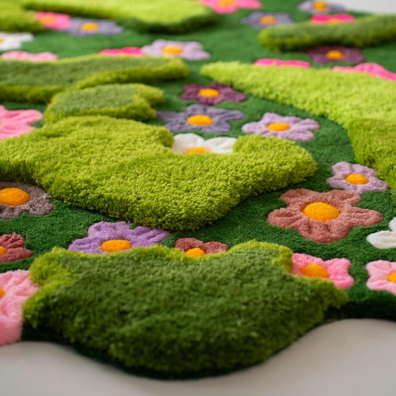 May include: A tufted rug featuring a deep green background with raised, textured patches of lime green and various small flowers in shades of pink, purple, white, and orange. The surface has a thick, plush pile with distinct, irregular shapes.