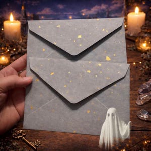 May include: Two stacked gray envelopes featuring a speckled gold foil pattern. The envelopes have triangular flaps and a textured surface. A small, white, semi-transparent ghost illustration is positioned at the bottom right corner.