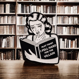 May include: Black and white pin of a crying woman reading a book. The book's cover reads "PLOT TWIST I FELL HARD FOR THE VILLAIN". The background is a bookshelf filled with books.