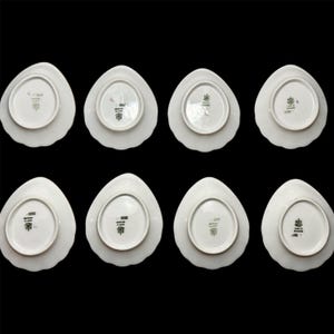 May include: Eight white porcelain plates with a unique, petal-like shape are arranged against a black background. The plates have a scalloped edge and a detailed emblem on the underside. The plates are likely for serving food.