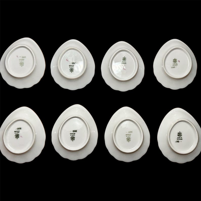 May include: Eight white porcelain plates with a unique, petal-like shape are arranged against a black background. The plates have a scalloped edge and a detailed emblem on the underside. The plates are likely for serving food.